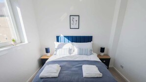 A neatly made bed with a blue headboard, white pillows, and two bedside lamps. - Derry House - spacious 3 bed house with driveway (Aberdeen)