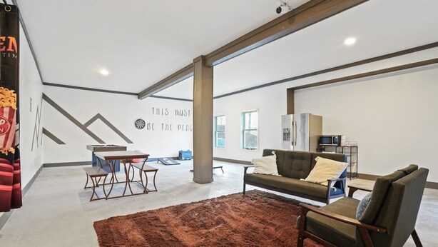 Ampla sala de estar com um tapete marrom grande, um sofá, uma poltrona e uma mesa de jantar. Há uma parede com um design geométrico e uma placa com a inscrição 'THIS MUST BE THE PLACE'.