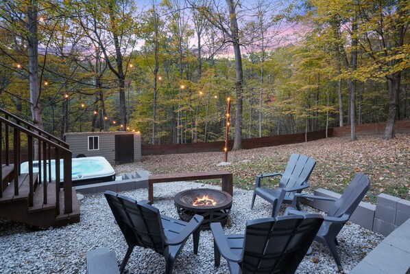 A deck with a hot tub, outdoor seating, and a fire pit surrounded by trees.