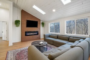 A modern living room with a fireplace, a large sofa, a coffee table, and a view of trees through the windows.