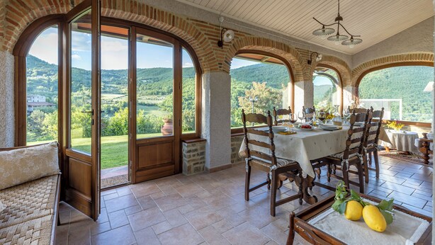 A dining area with a table set for four, large arched windows, and a view of a green landscape. - Private villa with pool, hot tub and garden in the Tuscan countryside (Castel Focognano)