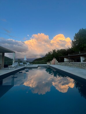 A swimming pool with lounge chairs and umbrellas, surrounded by stone walls and a view of the sky with clouds.