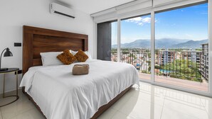 Ein geräumiges Schlafzimmer mit einem großen Bett, einem Holz-Kopfteil und Blick auf eine Stadtlandschaft durch raumhohe Fenster.