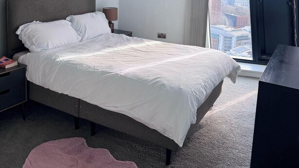 A modern bedroom with a large bed, a nightstand, a lamp, a painting, and a view of the city through a window. - City Centre - 2-bed High Rise Apartment - Sleeps 4 (Manchester)