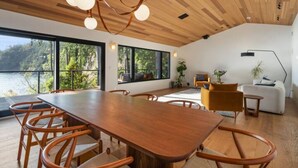 Un espace repas moderne avec une table et des chaises en bois, de grandes fenêtres donnant sur une zone boisée et un coin salon confortable avec un canapé et des fauteuils.