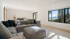Un salon moderne avec un canapé d’angle, un pouf et de grandes fenêtres donnant sur l’extérieur.