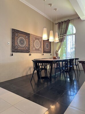 Un coin repas comprenant une table et des chaises, des éléments décoratifs muraux à motifs, des luminaires suspendus et une fenêtre avec des rideaux.