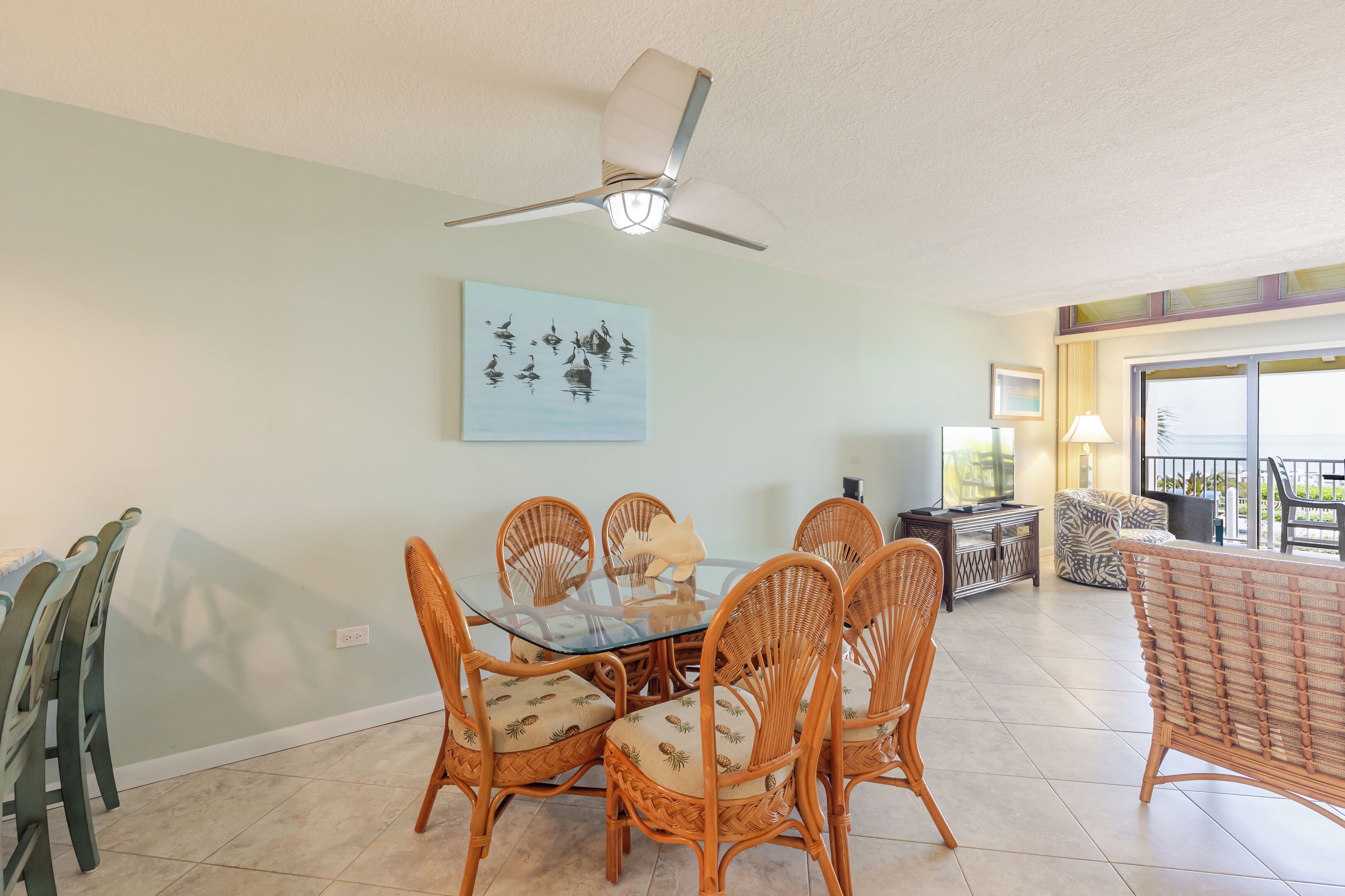 A dining area with a glass table surrounded by wicker chairs, a ceiling fan, and a painting on the wall.