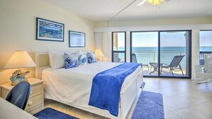 A bedroom with a bed, a nightstand, a lamp, a chair, a balcony with a view of the ocean, and a ceiling fan.