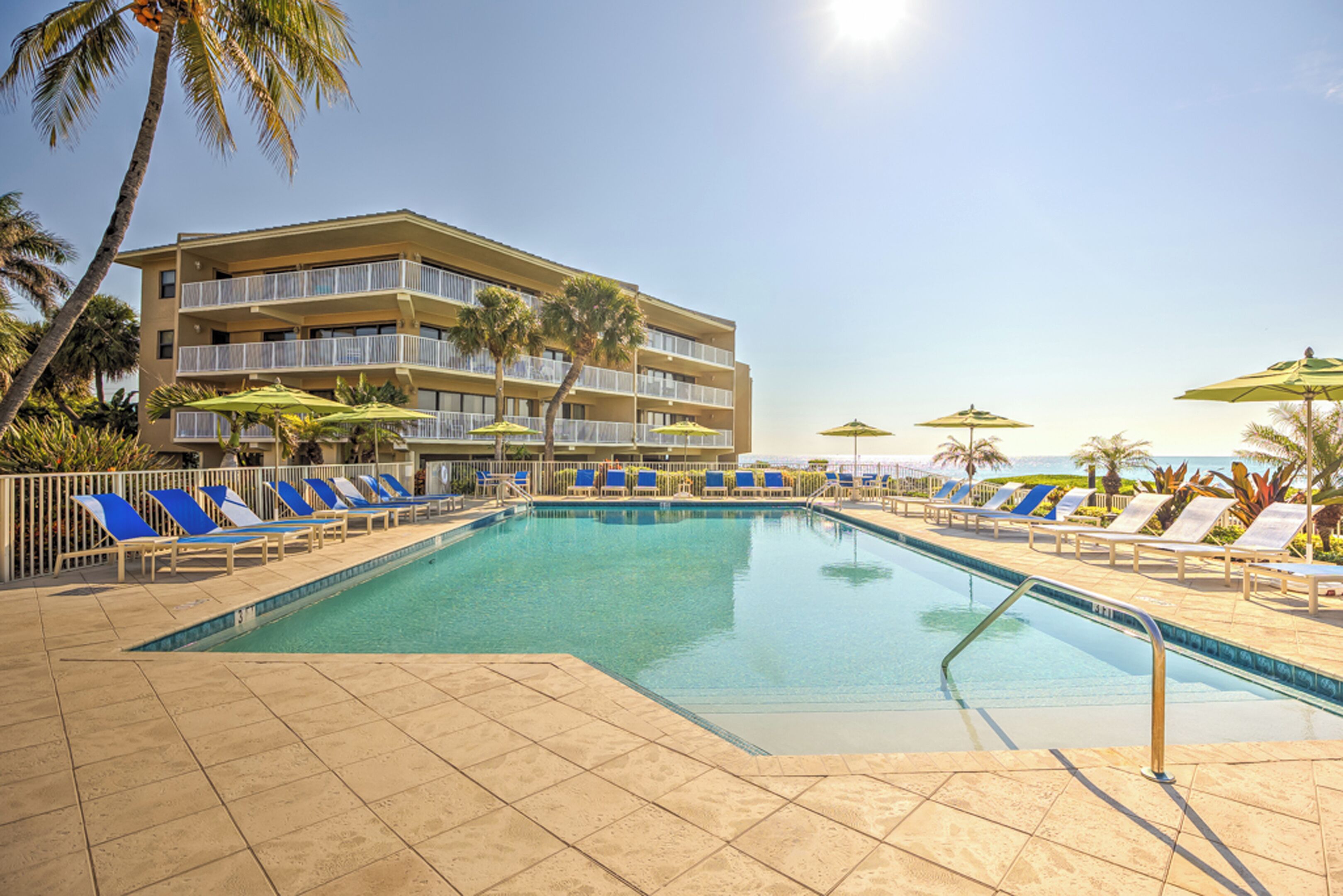 A pool area with blue lounge chairs and umbrellas, a multi-story building with balconies, and palm trees.