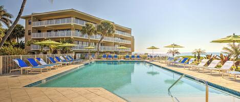 A pool area with blue lounge chairs and umbrellas, a multi-story building with balconies, and palm trees.