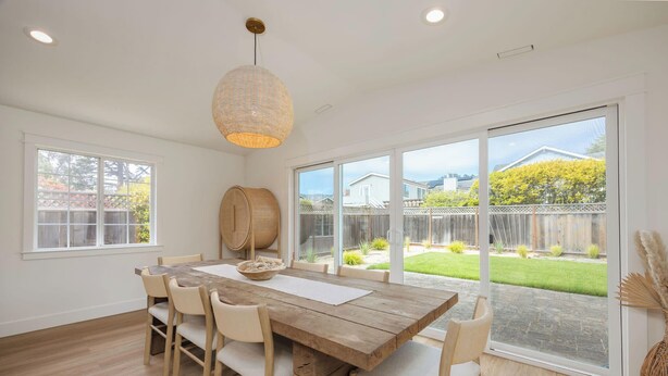 Un comedor con mesa y sillas de madera, ventanas amplias y una lámpara colgante.
