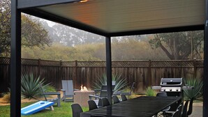 A covered patio with a grill, dining table, chairs, and a fire pit.