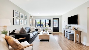 A modern living room with a sofa, coffee table, and TV. There are framed pictures on the wall and a window with a view of the outside. - King Beds in Safe Area • Game Room • Patio & Grill (Wichita)