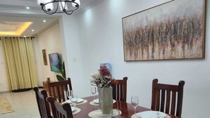 A dining area with a wooden table set for four, surrounded by chairs. A chandelier hangs from the ceiling, and a painting is on the wall.