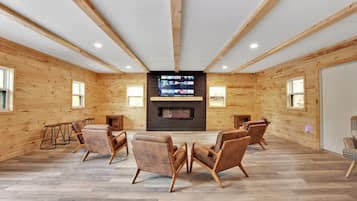 Ruime kamer met houten wanden, een groot televisiescherm en meerdere leren fauteuils rondom een centraal geplaatste open haard.