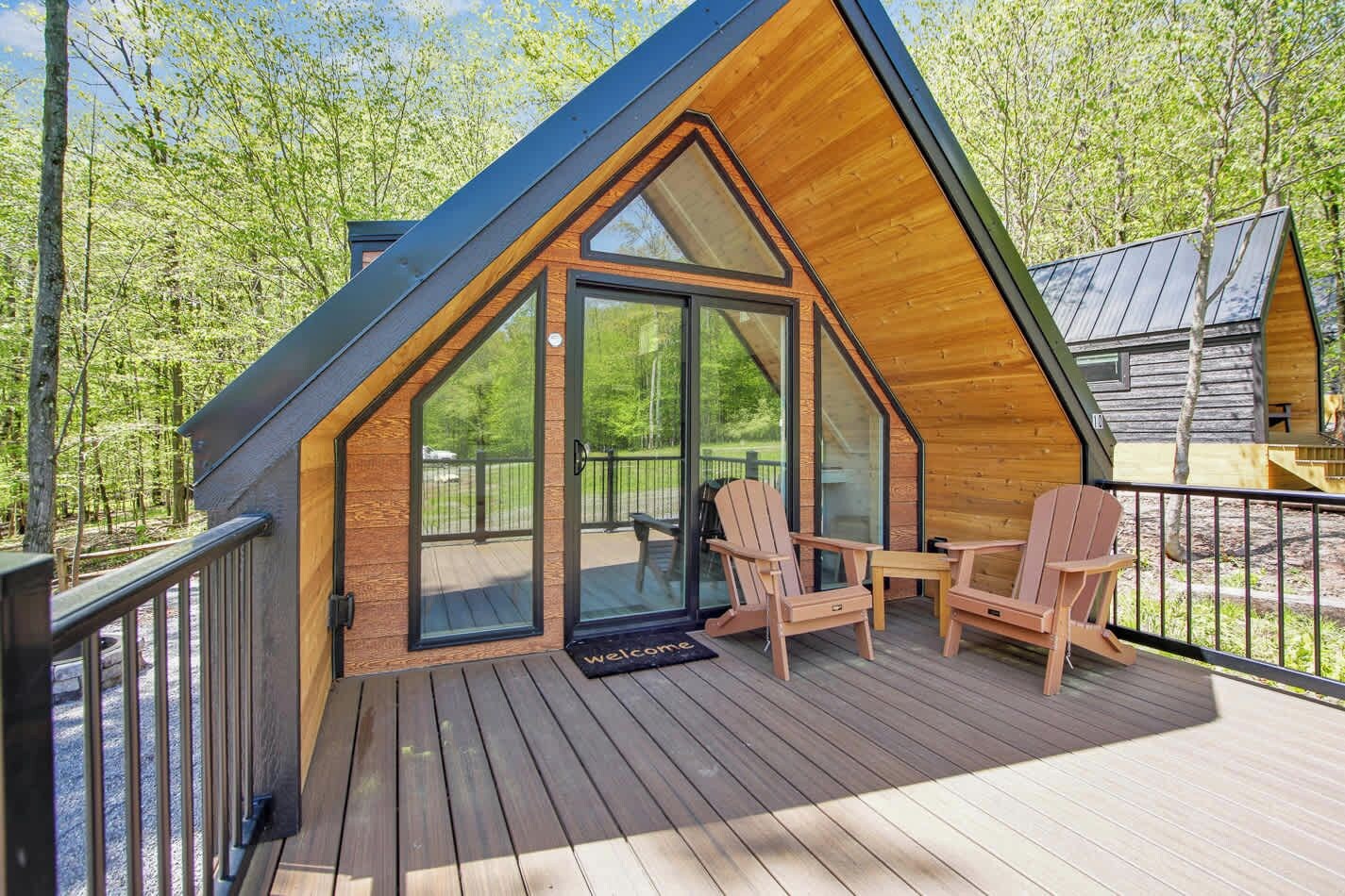 A cabin with a sloped roof, a deck with wooden chairs, and a glass door leading to an outdoor area.