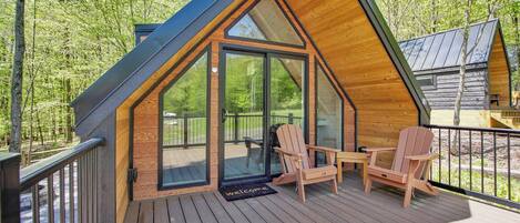 A cabin with a sloped roof, a deck with wooden chairs, and a glass door leading to an outdoor area.