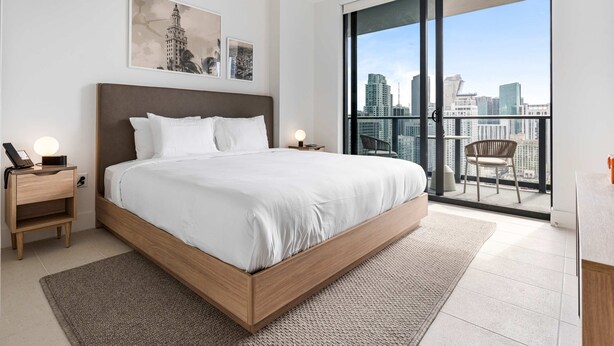 A modern bedroom with a large bed, a nightstand with a phone, and a view of a city skyline through a balcony.