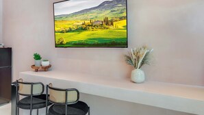 Ein modernes Wohnzimmer mit einem an der Wand montierten Flachbildfernseher, einem weißen Konsolentisch mit zwei Stühlen sowie dekorativen Elementen wie einer kleinen Topfpflanze und einer Vase mit Trockenblumen.