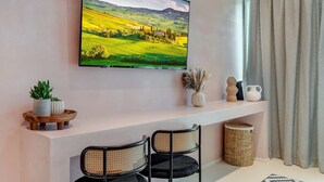 Ein Wohnzimmer mit einem an der Wand montierten Flachbildfernseher, einem weißen Konsolentisch, zwei schwarzen Stühlen mit Rattan-Sesseln, einer kleinen Topfpflanze, einer Vase mit Trockenblumen und einem Korbwäschekorb.