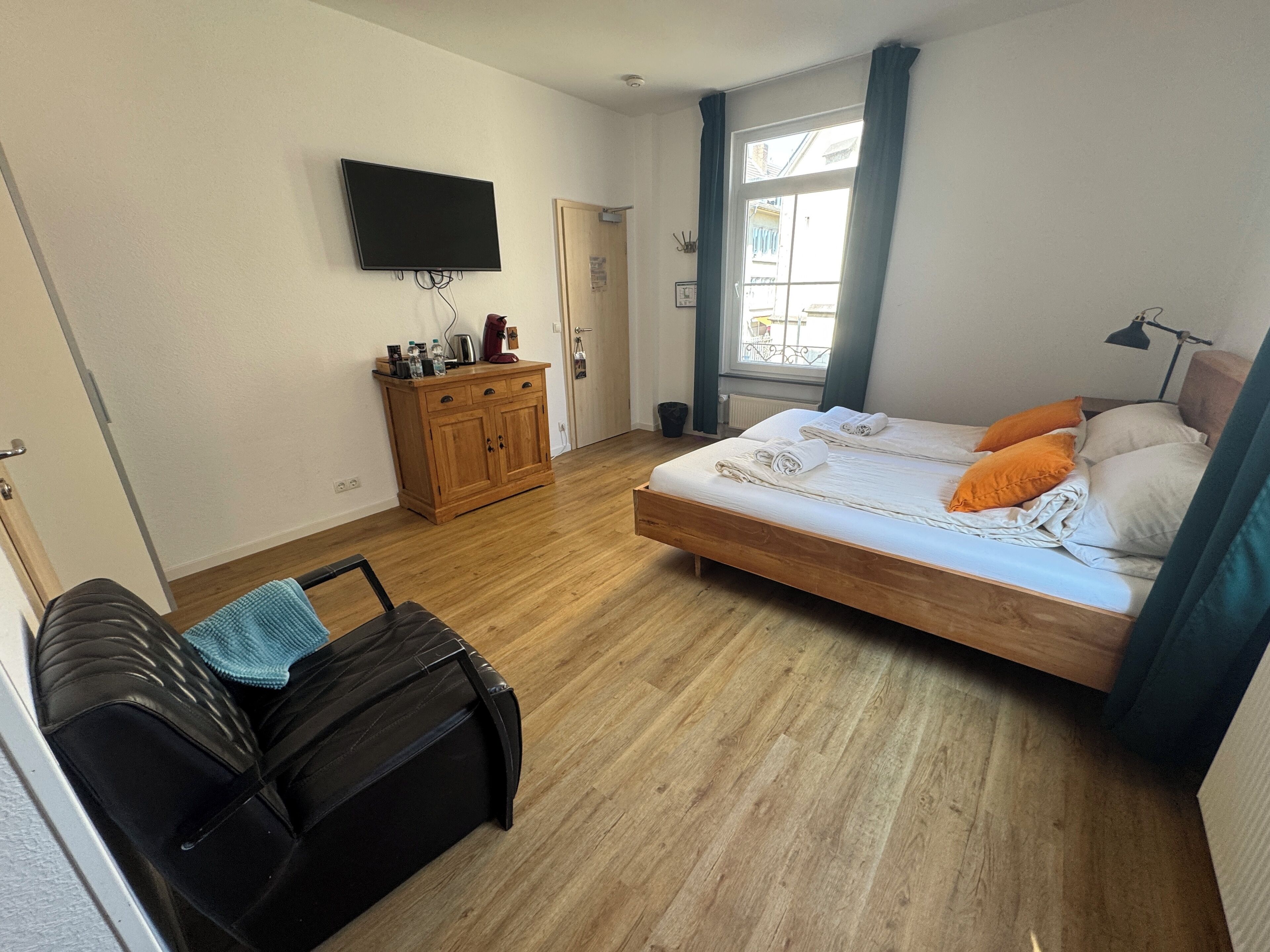 A bedroom with a bed, a TV mounted on the wall, a wooden dresser, and a window with curtains.