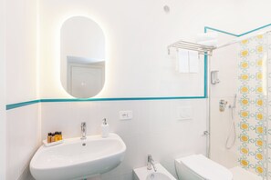 Un bagno con lavandino, specchio e water bianchi. È presente una zona doccia con rivestimento decorativo.