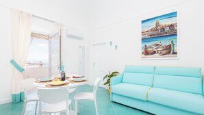 Una stanza luminosa con un divano color petrolio, sedie bianche, un tavolo da pranzo con piatti e un quadro raffigurante una veduta costiera.