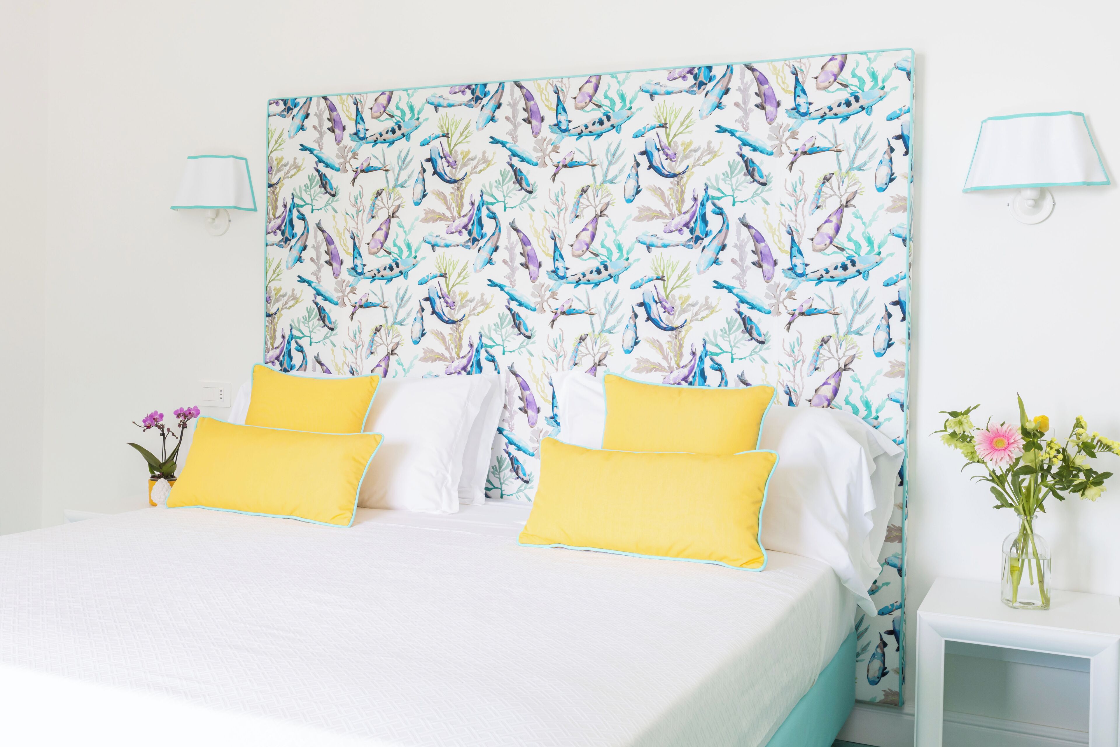A bed with white and yellow bedding, a floral headboard, and a nightstand with a vase of flowers.