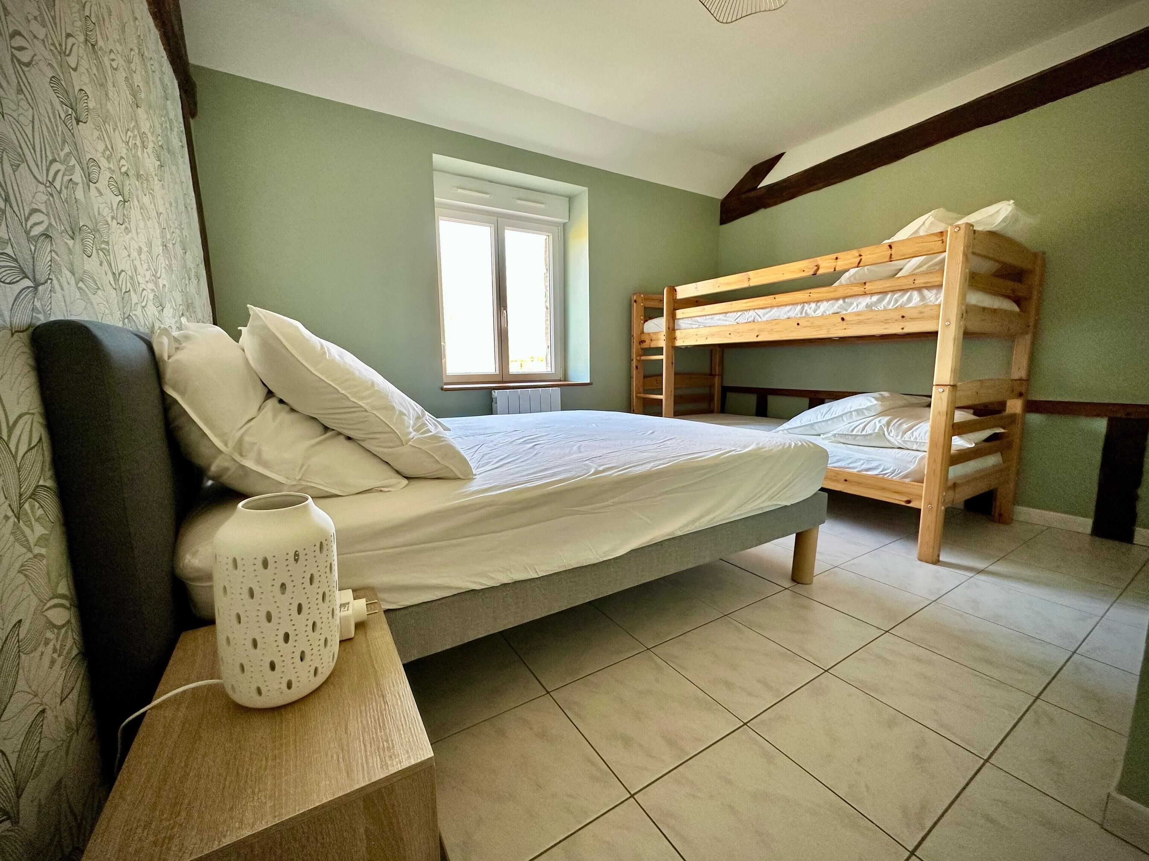 Una habitación con una cama nido, una cama individual, un ventanal y una mesita de noche con un jarrón blanco.