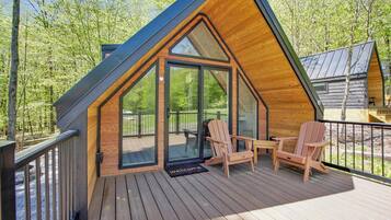 A cabin with a sloped roof, a deck with wooden chairs, and a glass door leading to an outdoor area.