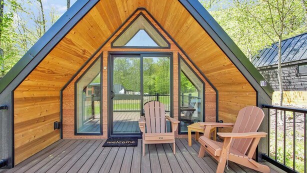 A wooden cabin with a sloped roof, a deck with two Adirondack chairs, and a glass door entrance. - Serenity Shack (Wellsboro)