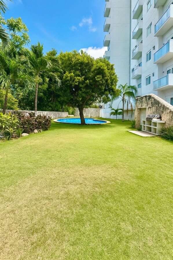 A well-maintained grassy area with a swimming pool, surrounded by trees and a multi-story building with balconies.