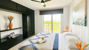 Ein modernes Schlafzimmer mit einem großen Bett, einem Deckenventilator, Blick auf Grünflächen und einem Bild an der Wand.