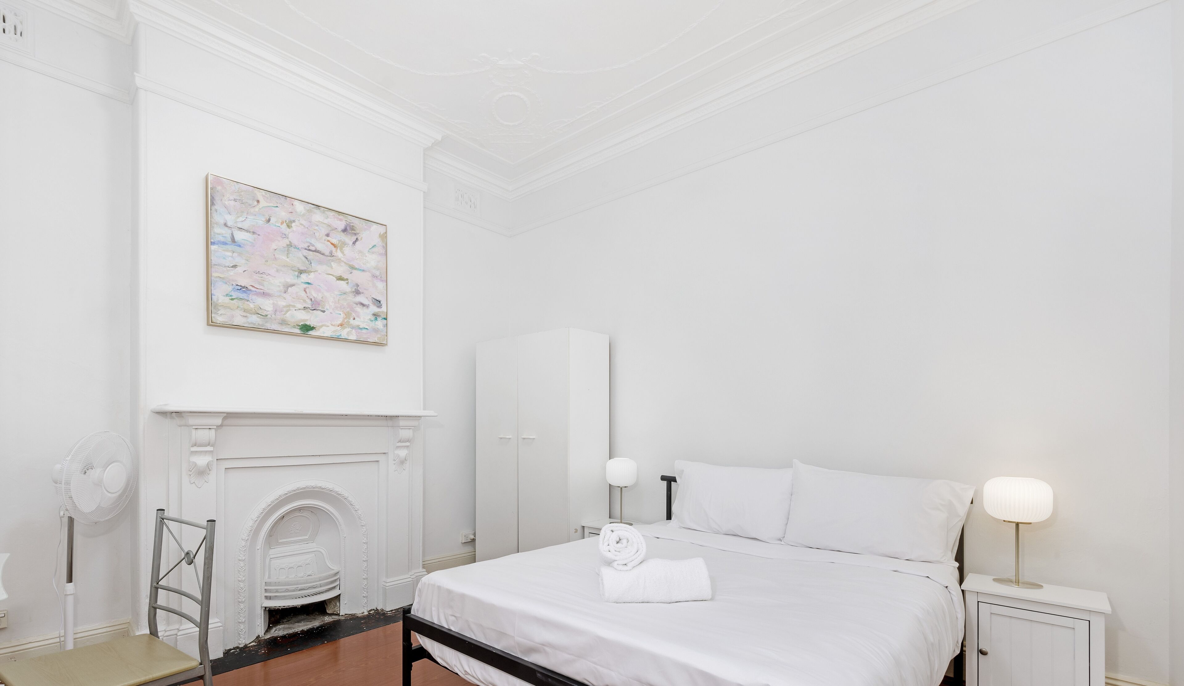Une pièce blanche avec un lit, une cheminée, un tableau et une table de chevet avec une lampe.