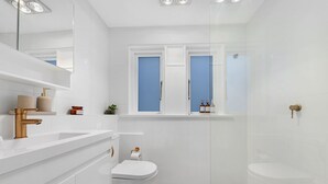 Un baño moderno con un lavamanos blanco, grifo dorado y un inodoro. Hay una ventana con vistas azules y un estante con artículos de aseo.