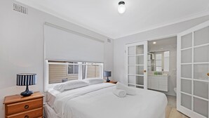 A bedroom with a bed, bedside tables, a dresser, a window, and a bathroom with a mirror and sink. - Kozyguru | Blacktown | Peaceful 3B House (Blacktown)