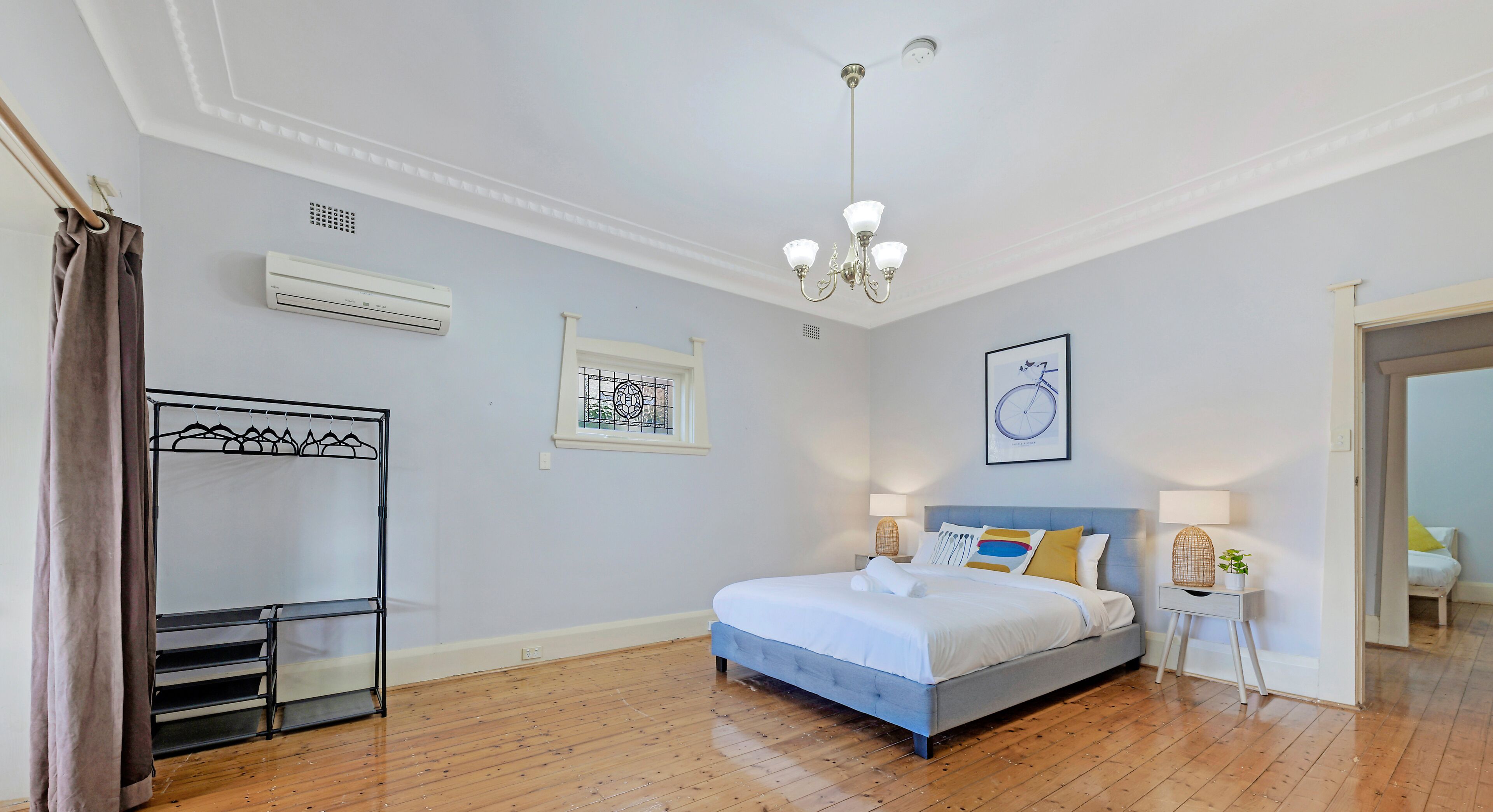 A bedroom with a bed, bedside lamps, a hanging light fixture, a window with a curtain, and a wall-mounted air conditioner.