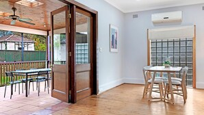 An open-plan living space with a wooden ceiling, a dining area with a table and chairs, and a view through a glass door to an outdoor patio. - Kozyguru | Campsie | Oldie-Goodie Look 4 Bed House (Campsie)