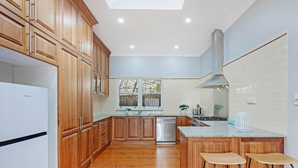 A modern kitchen with wooden cabinets, a stainless steel dishwasher, a granite countertop, and a large window. - Kozyguru | Campsie | Oldie-Goodie Look 4 Bed House (Campsie)