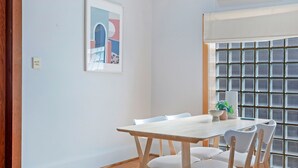 A dining area with a wooden table and chairs, a framed artwork on the wall, and an air conditioning unit above the window. - Kozyguru | Campsie | Oldie-Goodie Look 4 Bed House (Campsie)