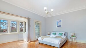 A bedroom with a bed, bedside lamps, a window with curtains, a chandelier, and a framed picture on the wall. - Kozyguru | Campsie | Oldie-Goodie Look 4 Bed House (Campsie)