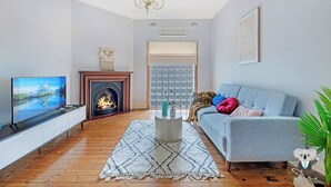 A living room with a television, fireplace, sofa, and a large window. - Kozyguru | Campsie | Oldie-Goodie Look 4 Bed House (Campsie)