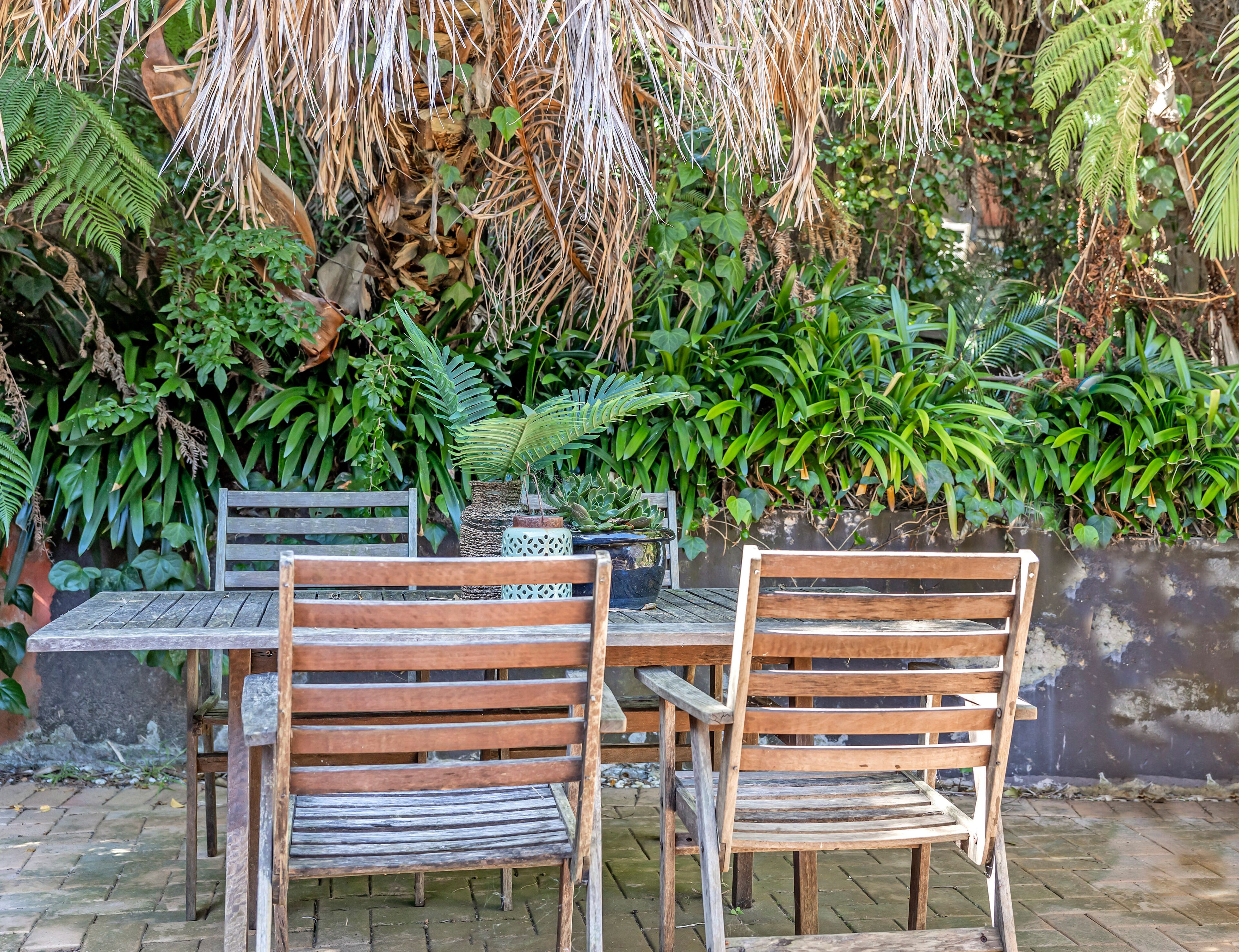 Ein Holztisch und zwei Stühle in einem Garten mit verschiedenen Pflanzen und einer Steinmauer im Hintergrund.