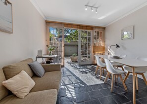 A modern living room with a sofa, dining table, and chairs. There is a sliding glass door leading to a patio area with a view of the outdoors. - Kozyguru | Glebe | Cozy 2 Bedroom Terrace House (Glebe)