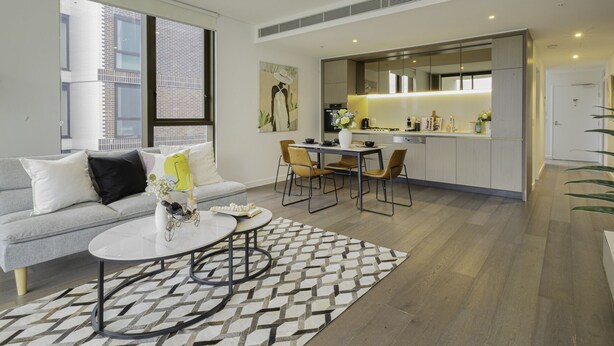 Un salon moderne avec un canapé, une table basse et un coin repas. On aperçoit une cuisine au fond.