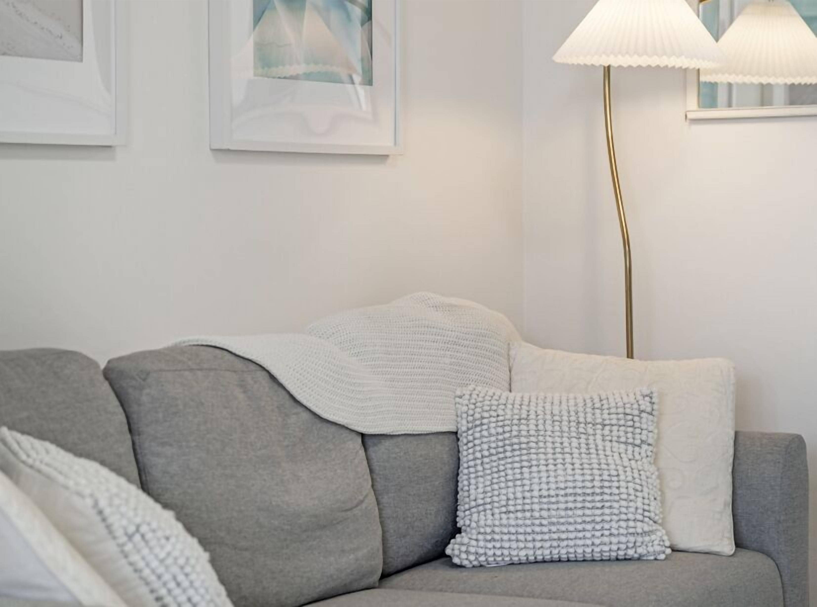 A grey sofa with a white throw blanket and a textured pillow, a floor lamp, and framed artwork on the wall.