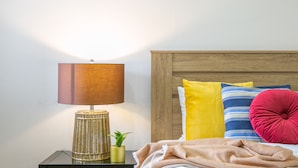 Een net opgemaakt bed met kleurrijke kussens, een bedlampje en een kleine plant in een potje op een nachtkastje.