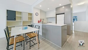 Une cuisine moderne avec des meubles gris, un plan de travail blanc et un îlot central intégré. On y trouve également un coin repas avec une table blanche et des chaises en osier.