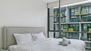 Une chambre moderne avec un grand lit, une table de chevet et une grande fenêtre donnant sur un immeuble doté de nombreux balcons.